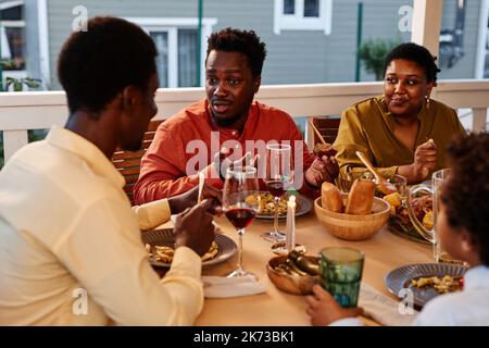 Porträt eines jungen schwarzen Mannes, der mit der Familie spricht, während er zusammen auf der Terrasse ein Abendessen genießt Stockfoto