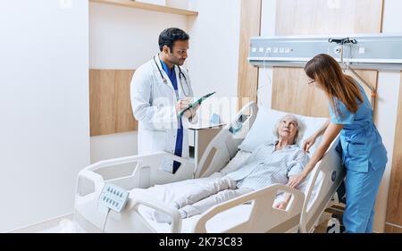 Bettseitberatung im modernen Krankenhaus. Ältere Frau, die auf dem Krankenhausbett auf der Station liegt, während sie sich mit einem freundlichen indischen Arzt und einer fürsorglichen Krankenschwester beraten hat Stockfoto