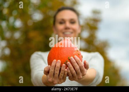 Hübsche Frau hält einen kleinen Kürbis in beiden Händen und zeigt ihn der Kamera und bietet ihn mit dem Fokus auf den Kürbis an Stockfoto