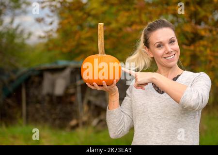 Eine hübsche Frau mittleren Alters, die mit einem großen Lächeln einen kleinen Kürbis in der Hand hält und vor einem herbstlichen Hintergrund mit dem Finger darauf zeigt Stockfoto