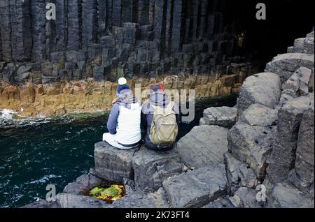 Zwei Menschen sitzen auf Felsen am Eingang zur Fingal's Cave auf der Isle of Staffa, Inner Hebrides, Schottland. Stockfoto