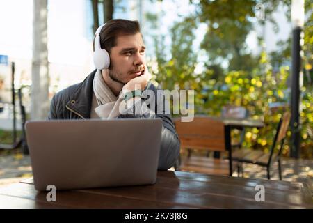 Nachdenklich erwachsener Mann arbeitet auf einem Laptop in Kopfhörern vor dem Hintergrund eines Herbstparks an einem Tisch in einem Café Stockfoto