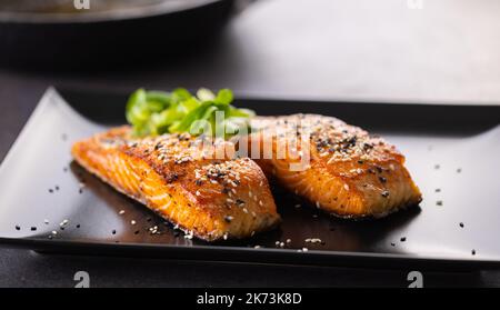 Zwei Lachsfilets, knusprig gebacken, mit Sesam auf einem schwarzen Teller bestreut. Stockfoto