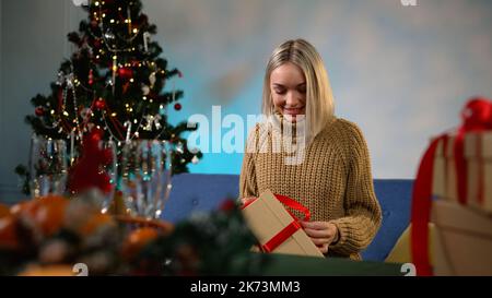 Das Mädchen öffnet ein Neujahrsgeschenk auf dem Sofa sitzend. Glückliches kleines Mädchen bindet ein rotes Band und öffnet ein Weihnachtsgeschenk zu Hause. Lächelnd nettes Mädchen g Stockfoto