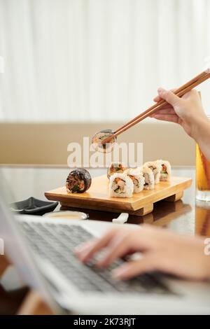 Junge Frau, die am Laptop arbeitet und Sushi isst, wenn sie zu Hause am Laptop arbeitet Stockfoto