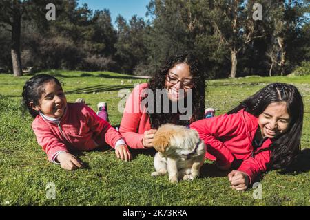 Junge Frau mit ihren beiden Töchtern, die auf dem Gras liegt und im Park mit einem goldenen Retriever-Welpen spielt Stockfoto