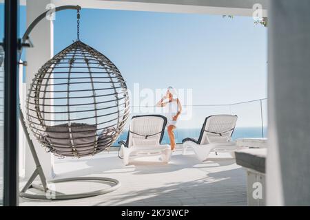 Nach der Dusche schöne Frau mit Haaren und Körper Handtuch wickeln stehen auf offener Terrasse mit Sonnenliegen, Hängesessel genießen Seeslandschaft und blau sauber Stockfoto