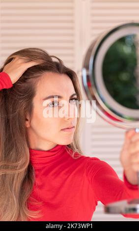 Vertikale Ansicht der jungen Frau, die an Alopezie leidet. Weiblicher Haarausfall.beunruhigte Frau, die ihren Haaransatz im Spiegel in rotem Outfit sieht Stockfoto