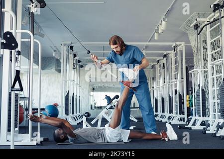 Männlicher Arzt in Uniform hilft Patienten mit Behinderung, sein Bein mit speziellen Sportgeräten zu strecken Stockfoto