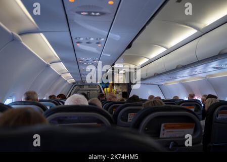 In der Passagierkabine eines easyJet Airbus A320 Airliner-Düsenflugzeugs mit Sitzen und Passagieren. Weibliches easyJet-Bodenpersonal, das sich mit der Crew begibt Stockfoto