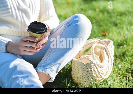 Eine Frau in einem weißen Pullover und einer blauen Jeans sitzt auf dem Gras und hält an einem sonnigen, hellen Tag ein Glas Kaffee in den Händen. Stockfoto