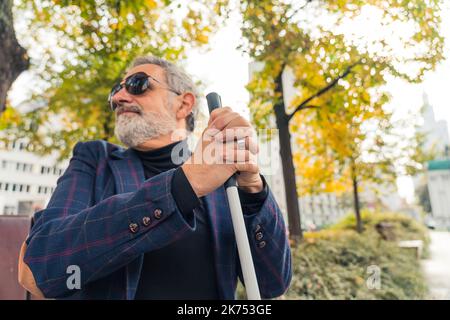 Blinder bärtiger grauhaariger reifer Mann mit dunkler Sonnenbrille, der einen Spazierstock hält und auf einer Bank im Park sitzt. Hochwertige Fotos Stockfoto