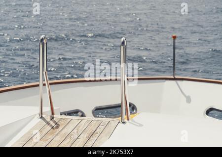 Blick vom Heck eines Yachthafens auf einen Teil des Bogens des Bootes mit selektivem Fokus auf ein verchromtes Leitergeländer und einen Holzboden; eine Leuchtfeuer und ein b Stockfoto