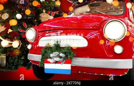 Das rote Neujahrsauto mit der luxemburgischen Flagge vor dem Hintergrund eines farbenfrohen Weihnachtshintergrunds Stockfoto