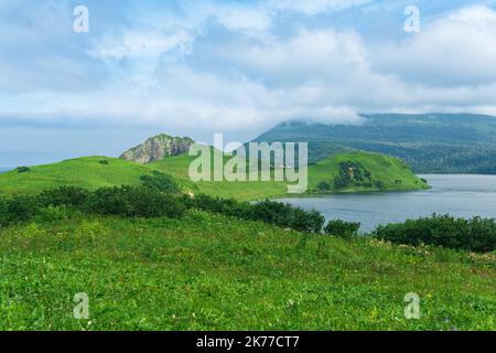 Natürliche Landschaft der Insel Kunashir mit grasbewachsenen Hügeln, vulkanischen Felsen, Vulkan in den Wolken und einem Tal mit einer Lagune Stockfoto