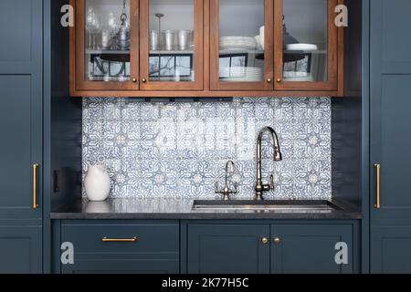 Ein Küchenwaschbecken mit einem schönen Muster gekachelten Backsplash mit einem Chrom-Wasserhahn, schwarzen Granit-Arbeitsplatten, und umgeben von blauen und Holzschränken. Stockfoto