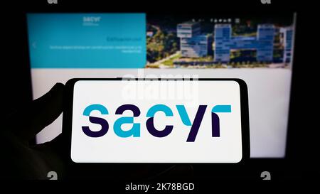 Person, die das Smartphone mit dem Logo des spanischen Infrastrukturunternehmens Sacyr S.A. auf dem Bildschirm vor der Website hält. Konzentrieren Sie sich auf die Telefonanzeige. Stockfoto