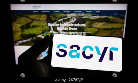 Person, die Mobiltelefon mit dem Logo des spanischen Infrastrukturunternehmens Sacyr S.A. auf dem Bildschirm vor der Business-Webseite hält. Konzentrieren Sie sich auf die Telefonanzeige. Stockfoto