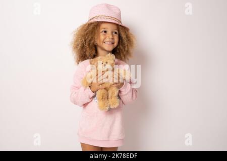 Schönes afroamerikanisches Kind, das Strohhut trägt und Teddybär umarmt und isoliert auf Weiß lächelt Stockfoto