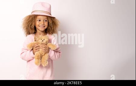 Wunderschönes afroamerikanisches Kind mit Strohhut, umarmt Teddybär und lächelt isoliert auf Weiß auf die Kamera Stockfoto