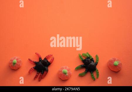 Hallowee: Gelee Bonbons in Form von riesigen schwarzen Spinnen mit grünen und roten Beinen und Kürbissen auf orangefarbenem Hintergrund. Speicherplatz kopieren. Stockfoto