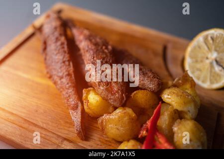 Gebratener Surmelettfisch mit Kartoffeln und Zitrone auf Holzbrett auf schwarzem Hintergrund Stockfoto