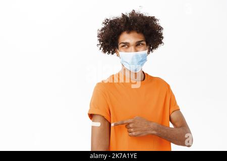 Der junge Mann impfte covid und zeigte mit Pflaster auf seinen Arm, er trägt eine medizinische Gesichtsmaske des Coronavirus, steht im orangen T-Shirt dagegen Stockfoto