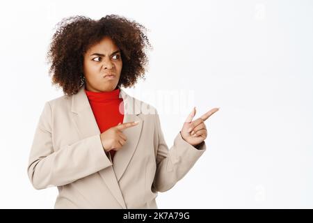 Verärgerte afroamerikanische Geschäftsfrau, die direkt auf leeren Raum zeigt und schaut, enttäuscht von smth, über weißem Hintergrund stehend Stockfoto