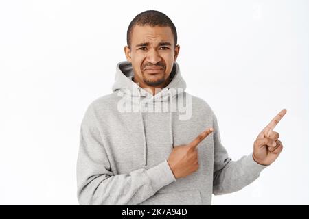 Weinender verstörter Kerl zeigt nach rechts, zeigt Werbung mit enttäuschtem Gesicht, steht in grauem Hoodie über weißem Hintergrund Stockfoto