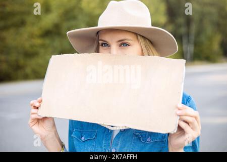 Porträt Nahaufnahme einer Frau, die hoffentlich auf das vorbeifahrende Auto im Wald wartet und leeres Pappplakat auf der Straße hält. Lady in Hut Flucht aus der Stadt durch Auto-Stop zu Stockfoto