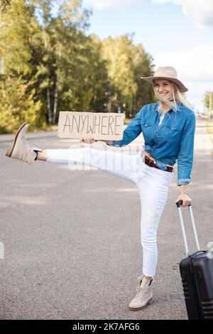 Freudige Frau mit Koffer versuchen, das vorbeifahrende Auto mit ausgestrecktem, erhöhtem Bein und einem Pappplakat auf einer leeren Autobahn zu stoppen. Lady in Hut Flucht aus der Stadt zu Stockfoto