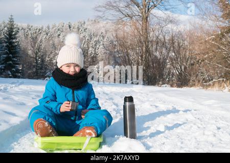 Junge sitzt auf dem Schlitten im winterverschneiten Land. Glückliches Kind Rodeln in der Natur. Kunststoffschlitten. Kind im Schlitten. Tee trinken. Stockfoto