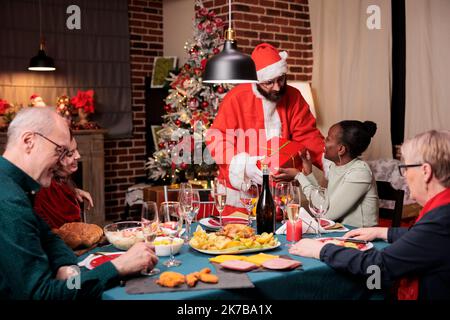 Familie tauscht weihnachtsgeschenke aus, afroamerikanische Frau bekommt Geschenk vom Mann im weihnachtsmann-Kostüm. Verschiedene Menschen feiern Weihnachten zu Hause Party, essen traditionelle Mahlzeit Stockfoto