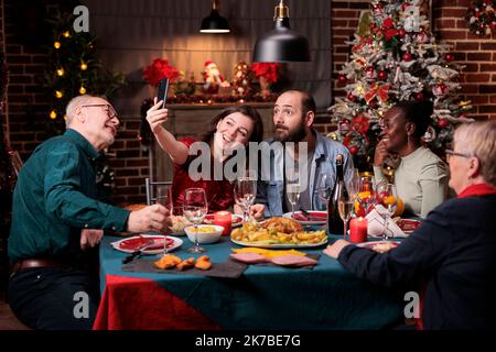 Frau, die ein Smartphone-Selfie der Weihnachtsfeier der Familie macht, Freunde porträtieren am festlichen Esstisch. Verschiedene Menschen treffen sich, feiern Weihnachten, essen traditionelle Mahlzeit Stockfoto