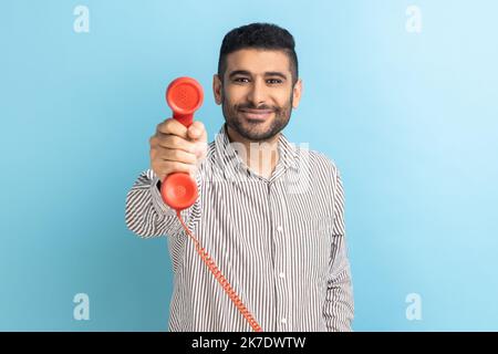 Nehmen Sie es an, und nehmen Sie den Anruf an. Mann, der das Telefon mit dem Hörer abgab und die Kamera ansah, lächelte freundlich, freundlich und hilfsbereit, das Callcenter trug ein gestreiftes Hemd. Innenaufnahme des Studios isoliert auf blauem Hintergrund. Stockfoto
