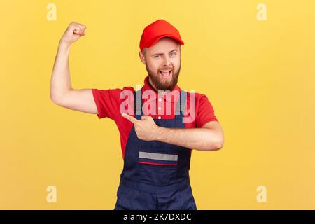 Porträt eines stolzen, starken Arbeiters in blauer Uniform und roter Mütze, der mit erhobenen Armen auf seinen Bizeps zeigt und seine Kraft zeigt. Innenaufnahme des Studios isoliert auf gelbem Hintergrund. Stockfoto