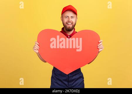 Porträt eines lächelnden romantischen Arbeiters mit blauen Overalls und roter Mütze mit großem roten Herzen, schnelle Geschenke während der Feiertage. Innenaufnahme des Studios isoliert auf gelbem Hintergrund. Stockfoto
