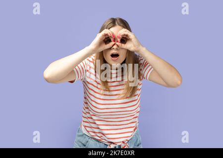 Porträt einer schockierten, erstaunten blonden Frau, die gestreiftes T-Shirt trägt und durch ein Fernglas schaut und einen schockierten Gesichtsausdruck hat. Innenaufnahme des Studios isoliert auf violettem Hintergrund. Stockfoto