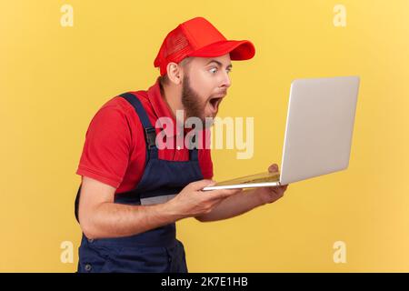 Seitenansicht eines äußerst schockierten Handwerkers, der seinen Laptop in den Händen hält und mit großen Augen auf das Notebook-Display blickt, das Uniform und rote Kappe trägt. Innenaufnahme des Studios isoliert auf gelbem Hintergrund. Stockfoto