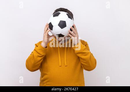 Anonymer unbekannter Mann, der sich hinter dem Fußball versteckt, ein Fußballfan bedeckt sein Gesicht während des Spiels und trägt einen Hoodie im urbanen Stil. Innenaufnahme des Studios isoliert auf weißem Hintergrund. Stockfoto