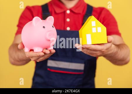 Porträt eines unbekannten anonymen Mannes mit blauer Uniform und rotem T-Shirt, der Sparschwein und Papierhaus zeigt und so Geld beim Kauf von Immobilien spart. Innenaufnahme des Studios isoliert auf gelbem Hintergrund. Stockfoto