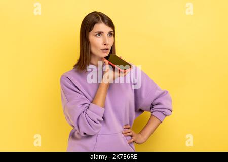 Porträt einer Frau, die mit einer digitalen sprach-App mit dem Smartphone spricht, Nachrichten aufzeichnet, Informationen im Internet sucht und einen violetten Hoodie trägt. Innenaufnahme des Studios isoliert auf gelbem Hintergrund. Stockfoto