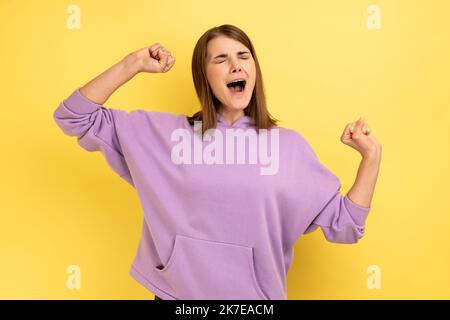 Porträt einer jungen, schlaflosen Frau, die die Hände aufhebt, sich müde fühlt, mit geschlossenen Augen steht und einen violetten Hoodie trägt. Innenaufnahme des Studios isoliert auf gelbem Hintergrund. Stockfoto