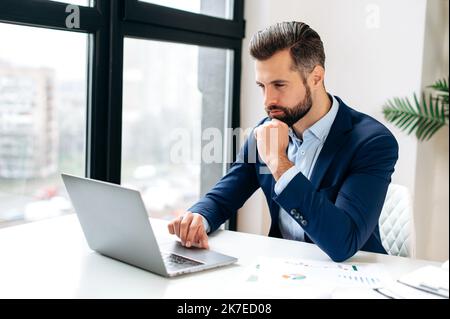 Fokussierter erfolgreicher selbstbewusster beschäftigter kaukasischer Geschäftsmann in einem formellen Anzug, sitzt am Arbeitsplatz, arbeitet an einem Projekt mit Laptop, schreibt Geschäftsstrategie, analysiert Informationen, im Internet surfen Stockfoto