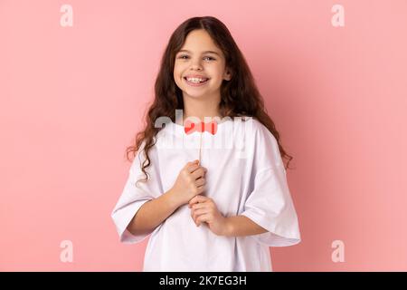 Porträt von zufriedenen kleinen Mädchen trägt weiße T-Shirt mit lustigen Papier Fliege, Spaß beim Spielen, trägt Maskerade Accessoire. Innenaufnahme des Studios isoliert auf rosa Hintergrund. Stockfoto