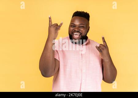 Porträt eines ungehorsamen Mannes in rosa Hemd mit Rock and Roll-Geste und aufgeregt schreiend, Spaß habend, genießen schwere Musik. Innenaufnahme des Studios isoliert auf gelbem Hintergrund. Stockfoto