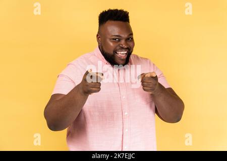 Porträt eines fröhlichen Mannes in einem rosa Hemd, der auf die Kamera zeigt und mit einem charmanten, toothy Lächeln, dem Finger und der Wahl aussieht. Innenaufnahme des Studios isoliert auf gelbem Hintergrund. Stockfoto