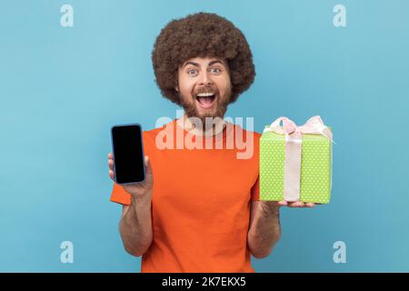 Porträt eines aufgeregt staunenden Mannes mit Afro-Frisur in orangefarbenem T-Shirt, der mit einer Präsenzbox steht und das Handy mit leerem Display zeigt. Innenaufnahme des Studios isoliert auf blauem Hintergrund. Stockfoto