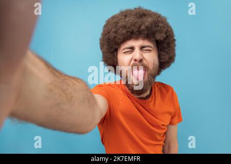 Porträt eines kindisch albernen Mannes mit Afro-Frisur in orangefarbenem T-Shirt, der Selfie macht oder Livestream macht, die Zunge zeigt, Sichtfoto. Innenaufnahme des Studios isoliert auf blauem Hintergrund. Stockfoto