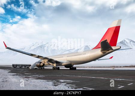 Breitkörper-Passagierflugzeug mit Treppe auf dem Flughafenvorfeld auf dem Hintergrund der hoch gelegenen schneebedeckten Berge. Shuttle-Bus in der Nähe des Flugzeugs Stockfoto